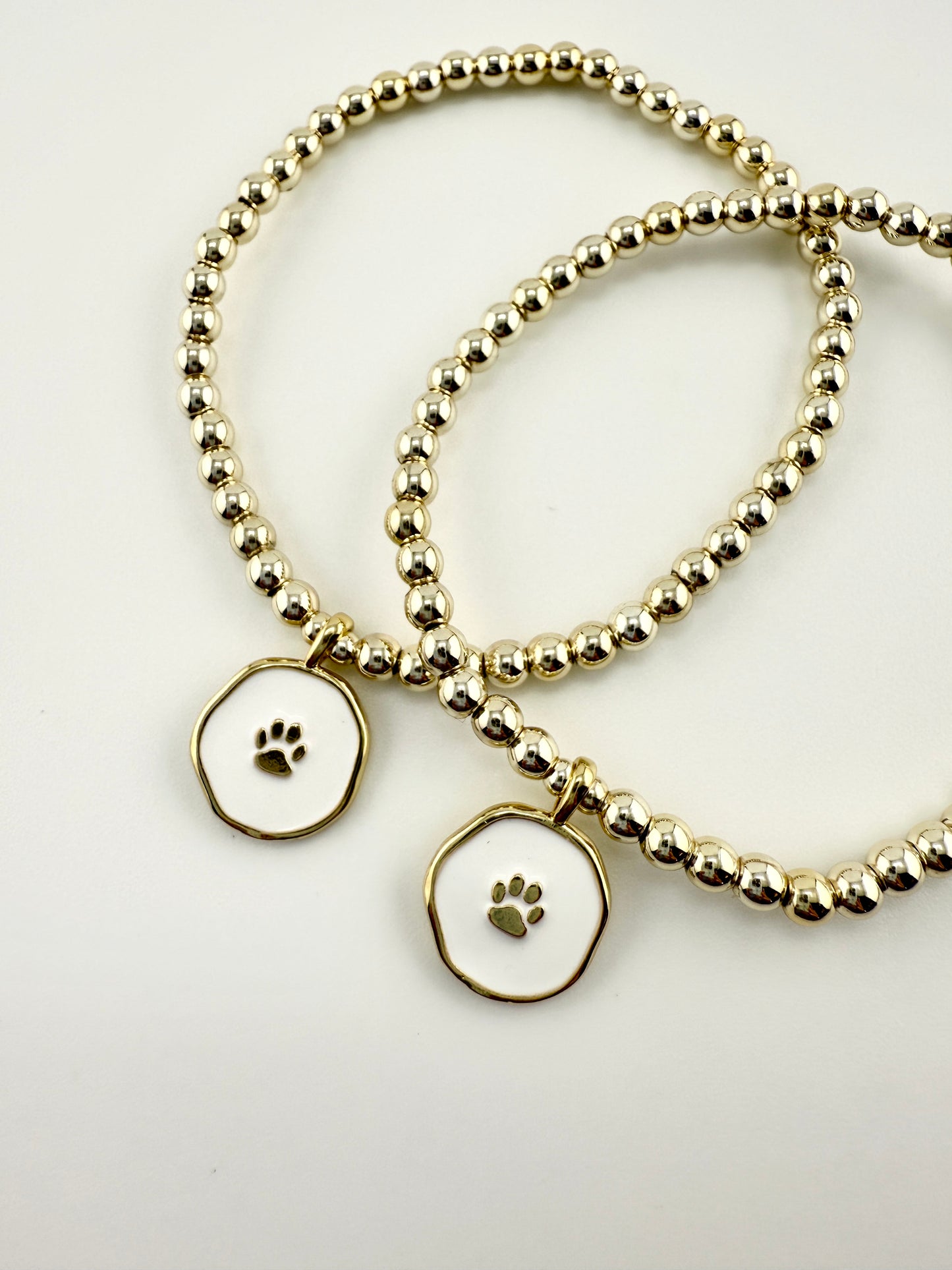 Two gold beaded bracelets with paw print charms on a light gray background